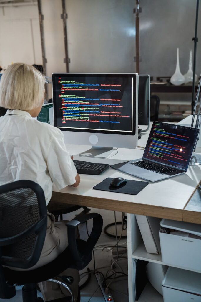 back view of a blond woman in an office looking at two computer screens with multicoloured code
