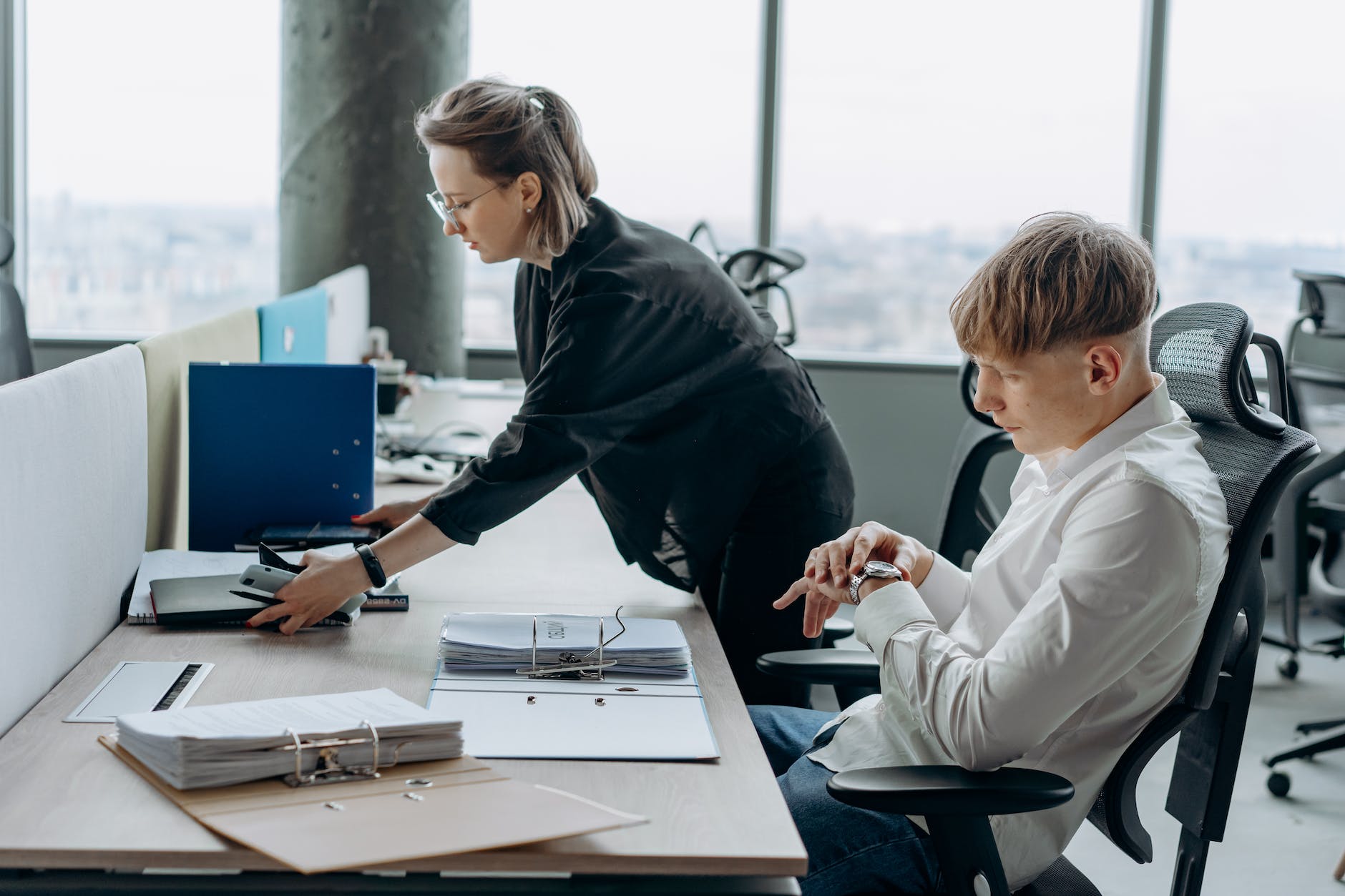 young man sitting in white long sleeve shirt looking on his watch beside his coworker inside an office